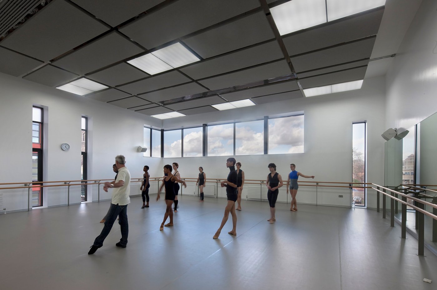 A ballet dance class inside the bright Jerwood Dance House DanceEast studio with students practicing at the barre, with large windows streaming in daylight, and a ceiling with additional lighting panels.