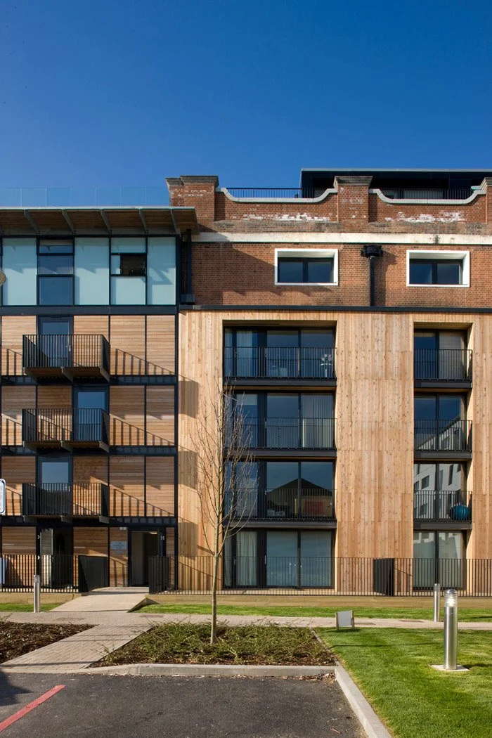 Modern multi-story apartment view of The Pump House with wooden and brick exterior, balconies, glass windows, and a landscaped front yard with a small tree, sidewalk, and parking lot.