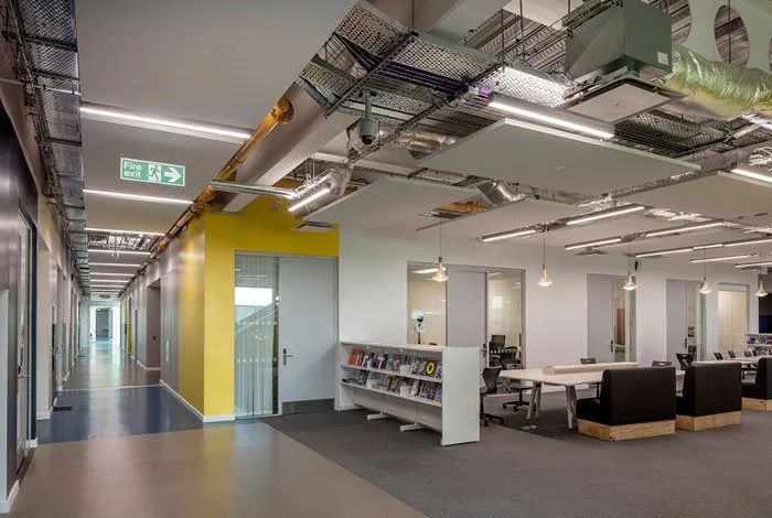 Modern office space with an open layout, featuring a white bookshelf, a row of black chairs, and hanging light fixtures, with exposed ceilings and a corridor with a yellow accent wall.