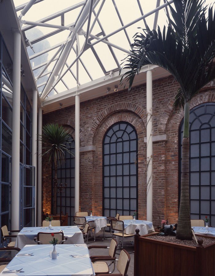 An internal view of White Cloth Hall with and indoor restaurant with brick walls, large arched windows, and a glass ceiling, decorated with potted plants and set tables with white tablecloths.