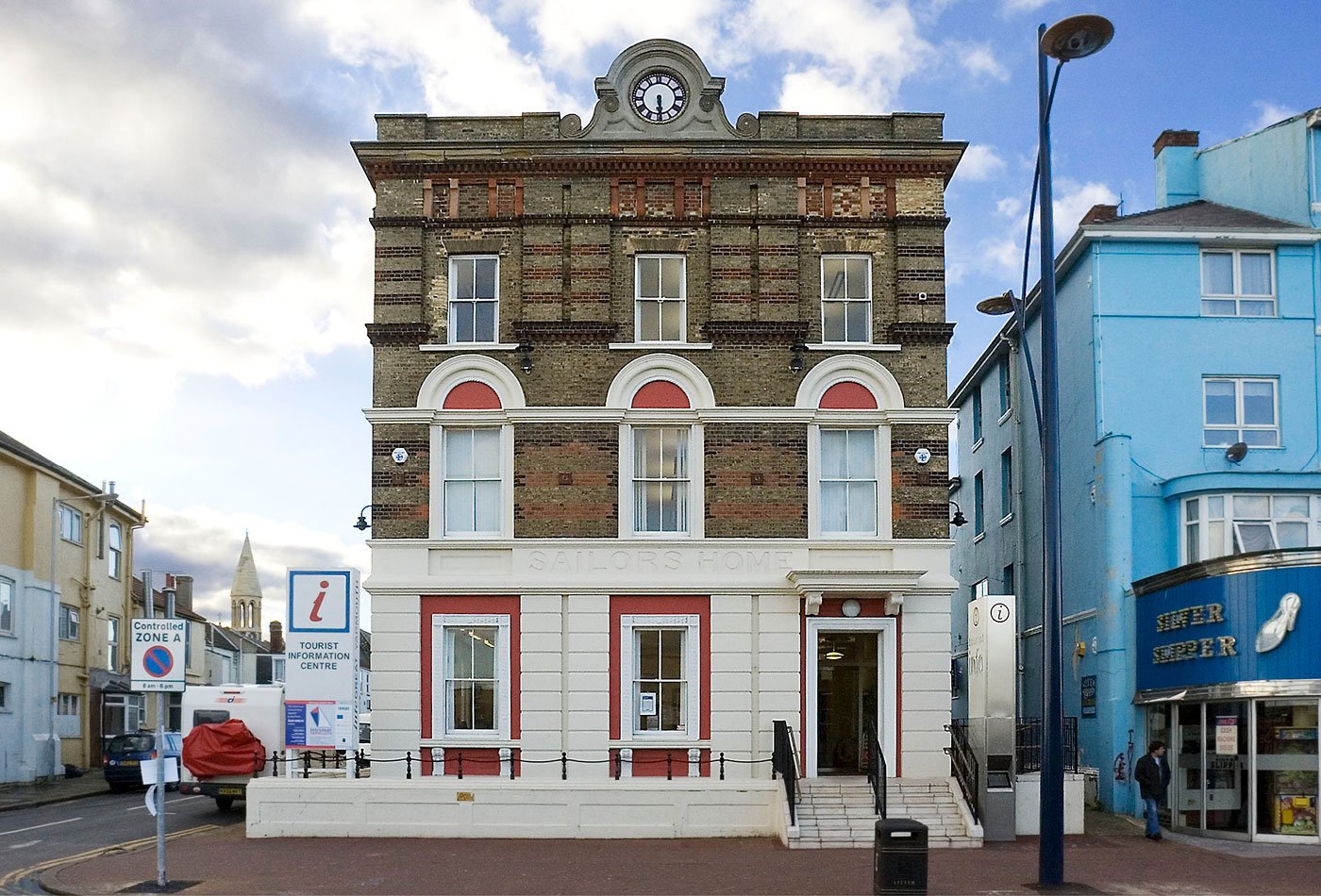 A square-on symmetrical view of the front of the Tourist Information Centre with historic brick work against the white lower render with 'Sailors Home' original signage and clock face, situated on a street corner with a bordering shop and signs.