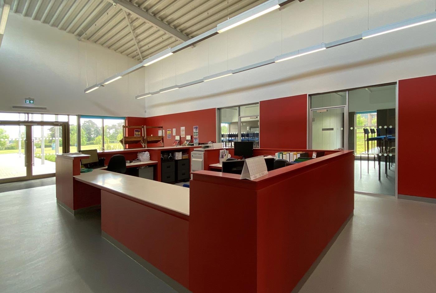 Empty reception area with red and white counter, office chairs, books, and glass doors leading outside, with bright natural light.