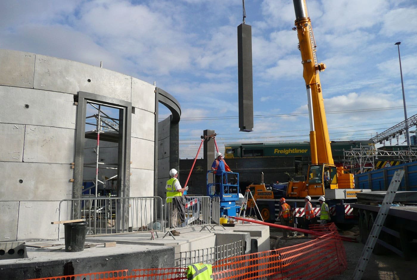 Construction workers are operating a crane to lift a concrete panel onto a building at a construction site, with workers wearing safety vests and helmets, surrounded by scaffolding, fencing, and construction equipment.