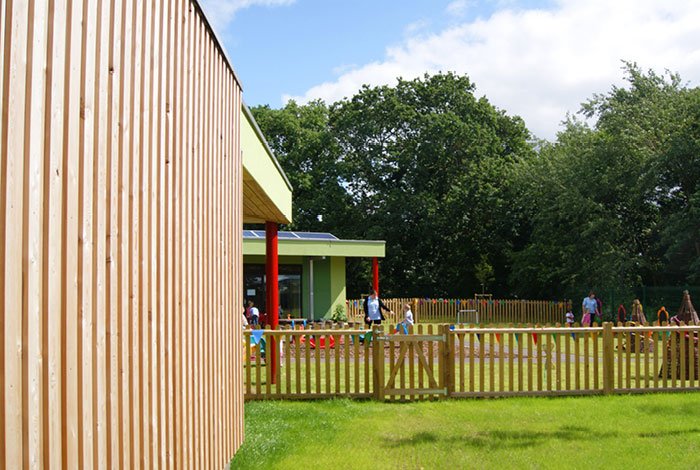 A side view of the children playing in a fenced outdoor area near a green building with red columns, surrounded by trees.