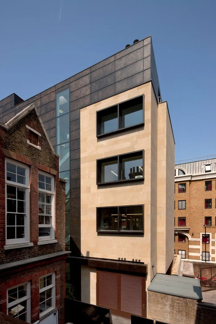 Modern multi-story building with large black-framed windows, beige stone facade, and black metal roof, situated between older brick buildings in an urban area under a clear blue sky.