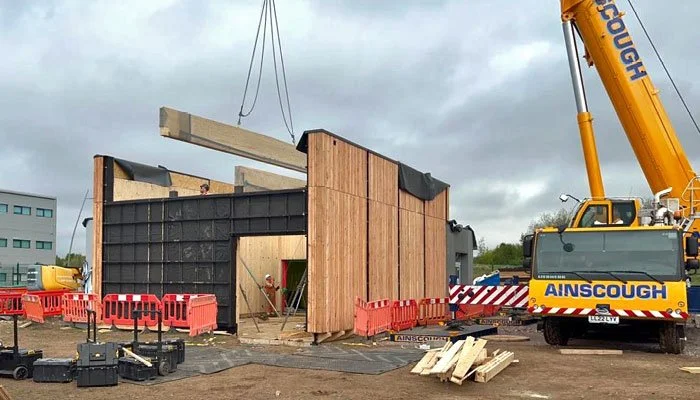 Construction site with a large yellow crane lifting a wooden beam, partially built wooden structure, and orange safety barriers.