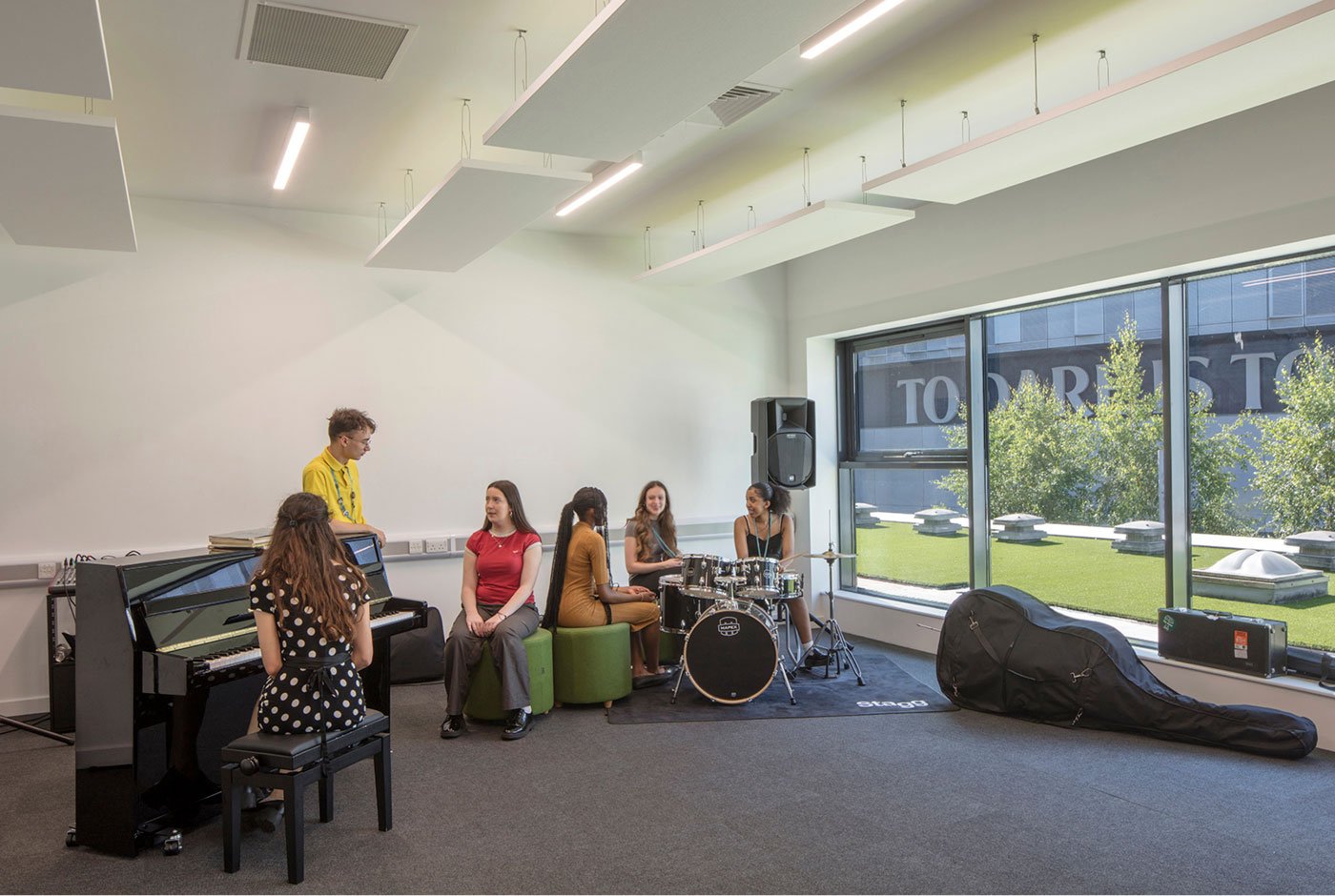 Group of young people practicing music in a well-lit room with large windows, featuring a piano, drums, and a guitar case.