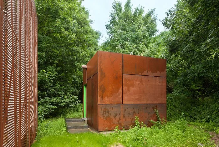 An isolated small rust-coloured metal cube building surrounded by green trees and grass, with a set of concrete steps leading to a green door.