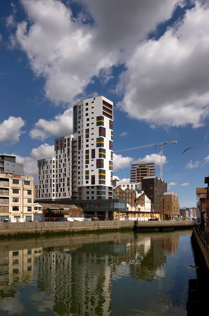 Modern high-rise building with colorful rectangular accents in an urban setting next to water, with partly cloudy sky and construction crane in the background.