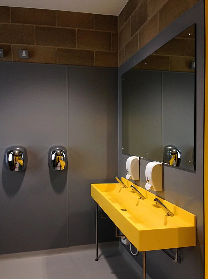 A modern bathroom with a yellow rectangular sink, two white soap dispensers, two mirror fixtures, and two chrome hand dryers on a dark gray wall, with a large mirror reflecting the scene.