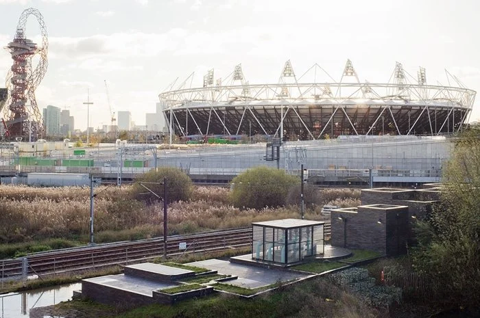 A large sports stadium with an open roof, surrounded by urban buildings, with a railway track and some small structures in the foreground.