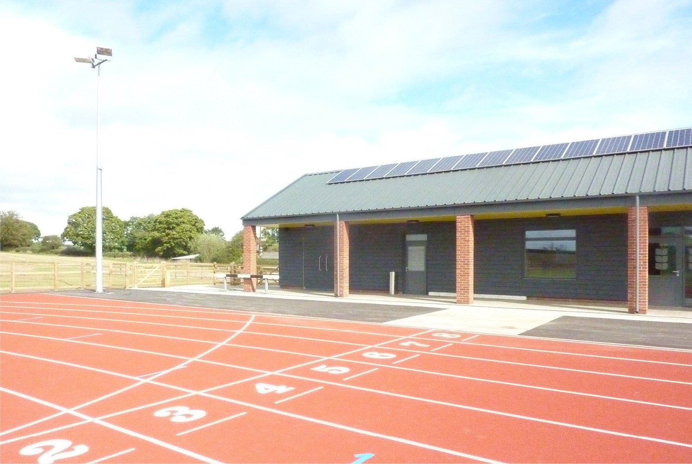 Outdoor sports track with numbered lanes, adjacent to a building with solar panels on the roof, green trees in the background, and a clear blue sky.