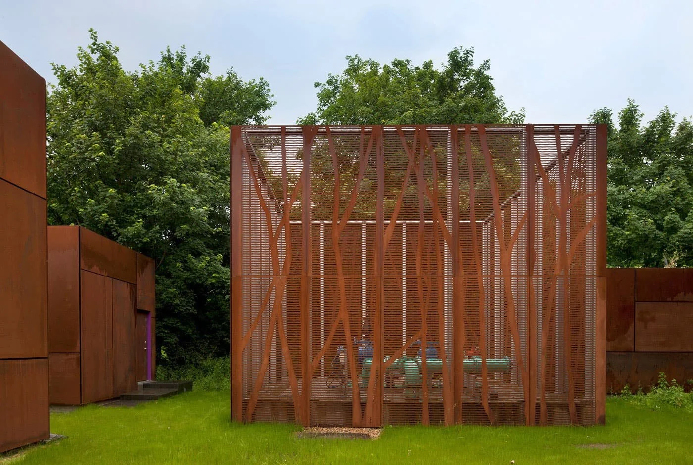A closeup of one of Old Ford Pumping Station outdoor structures with a rust-coloured caging that surrounds industrial pipe terminals, tree-branch-shaped lattice design, surrounded by green grass and trees.