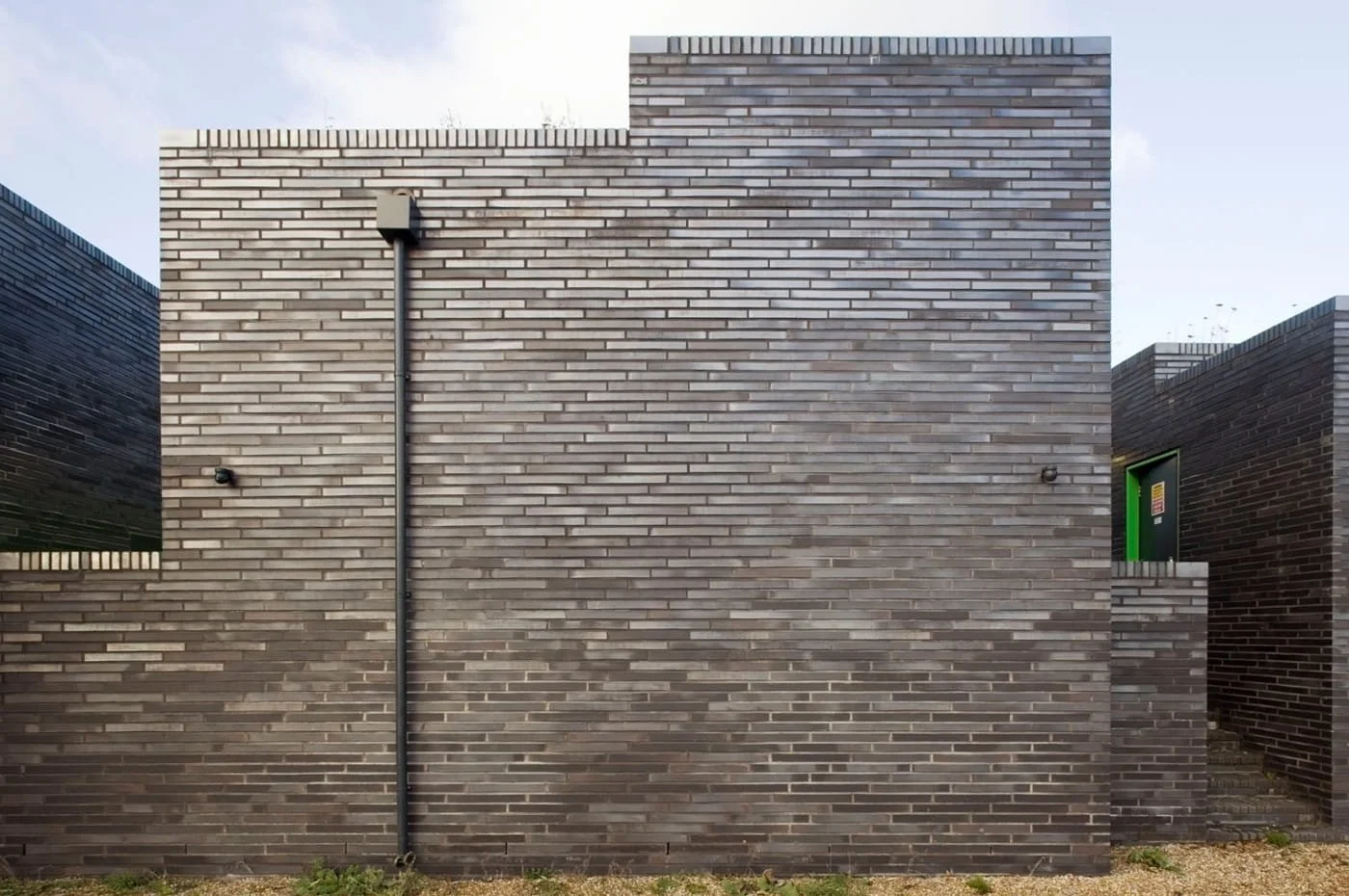 A modern building with a gray brick facade, a black conduit running vertically, and a small green door on the side.