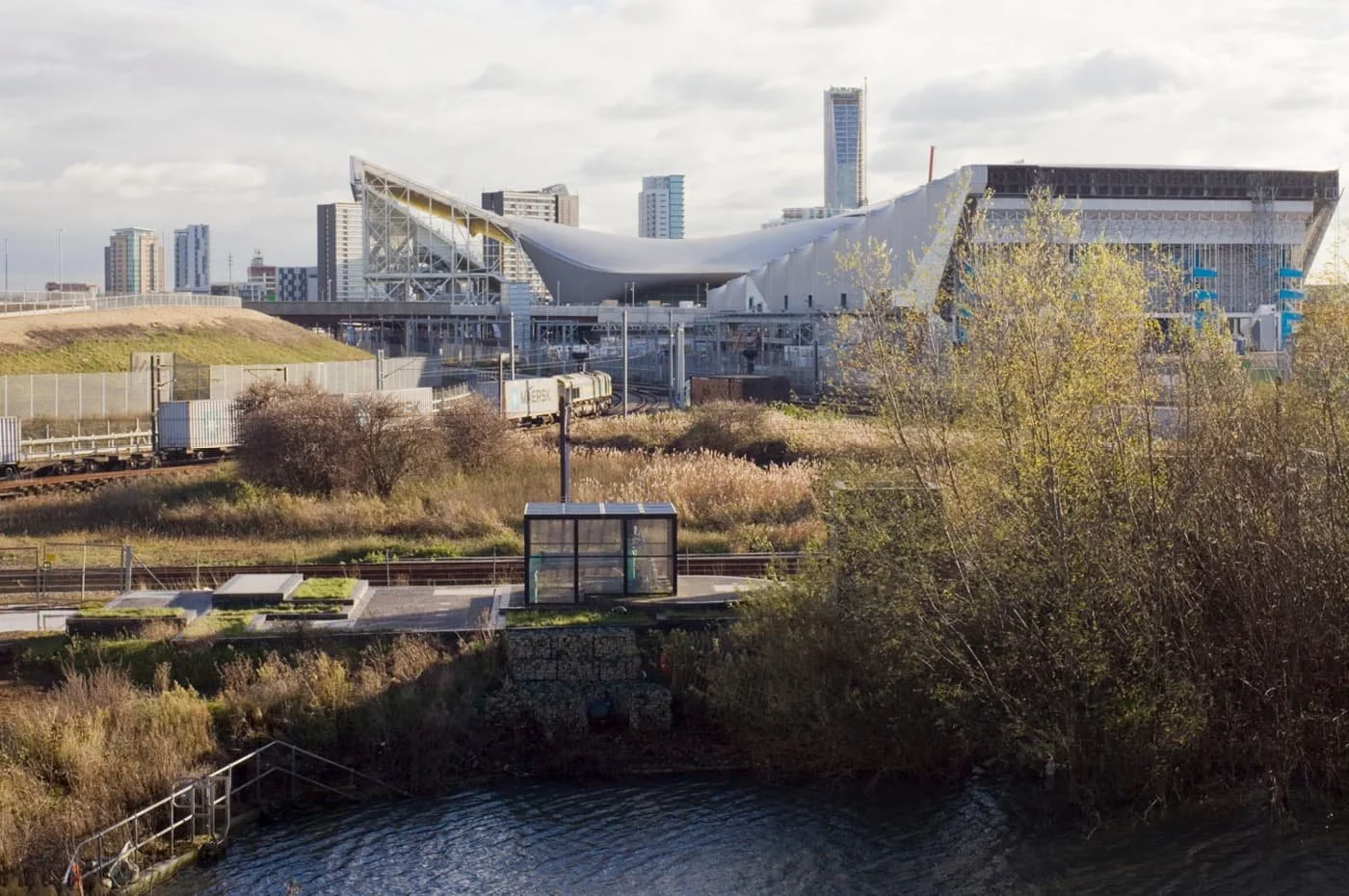 Cityscape with modern architecture building, train tracks, greenery, and a body of water in the foreground.