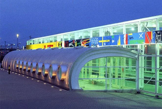 Tottenham Hale Station with a curved metallic entrance, glass walls, and colourful abstract art along the top, illuminated against a dusk sky.