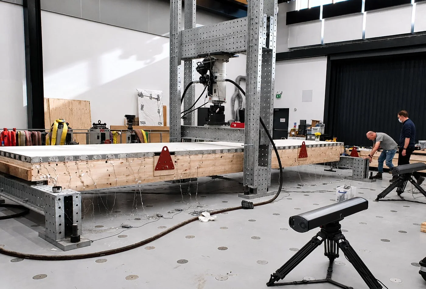 A large experimental setup with a robotic arm on a metal frame above a wooden platform, in a spacious industrial or research laboratory. Two men are working on the right side of the setup, one is crouching and another is standing nearby, both wearing masks.