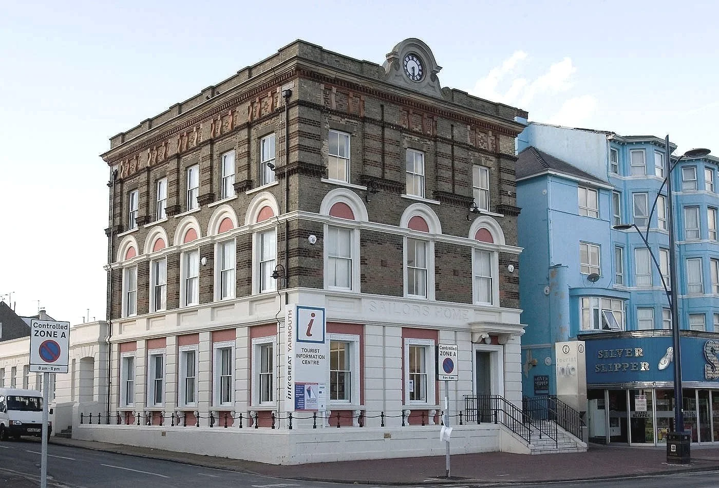 The historic multi-story Tourist Information Centre building in Great Yarmouth with a clock on top, featuring large windows and decorative architectural details, next to a blue building with white trim, street signs, and a pedestrian sidewalk.