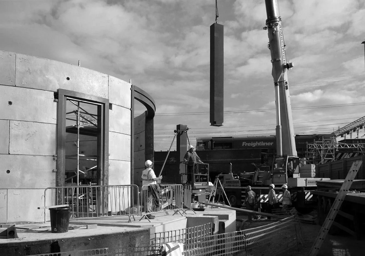 Construction site with workers in safety helmets and vests, crane lifting a large rectangular object, and a partially built curved concrete structure.