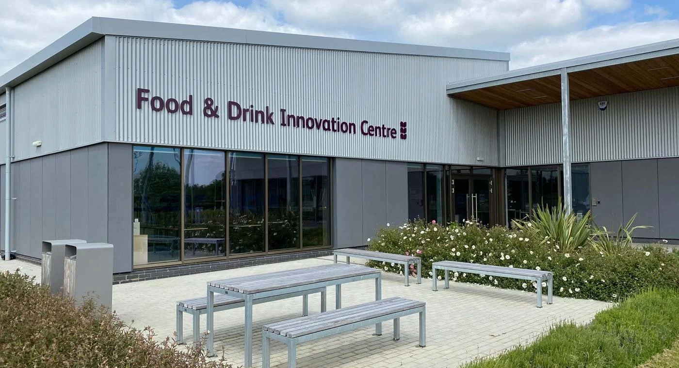 Exterior of a modern building labeled 'Food & Drink Innovation Centre' with benches and landscaped bushes in front.