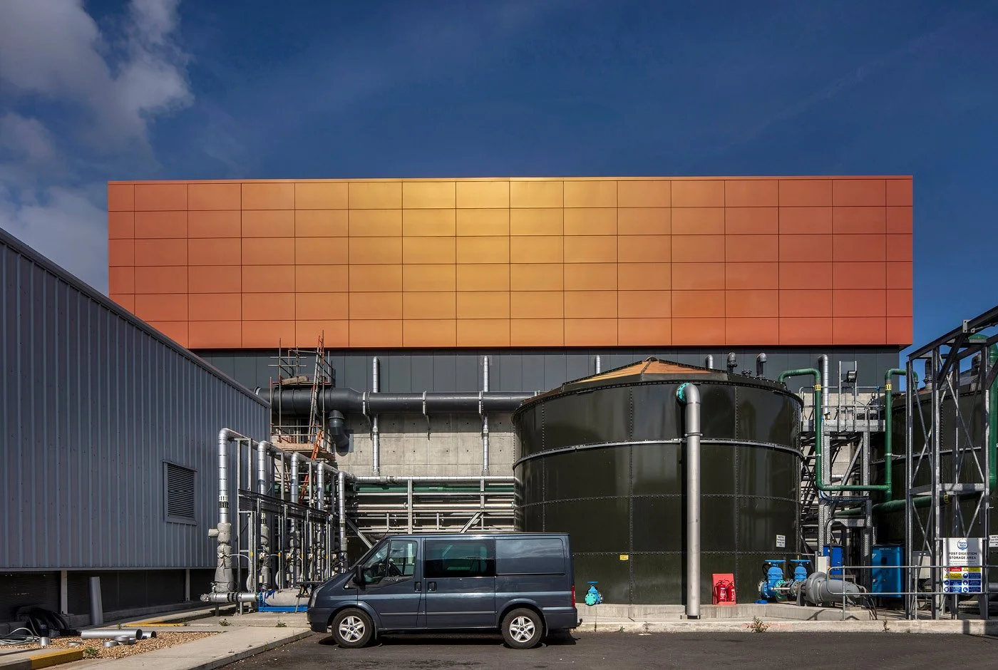 Industrial building with orange upper facade, black water tank, pipes, and a dark gray van parked in front.