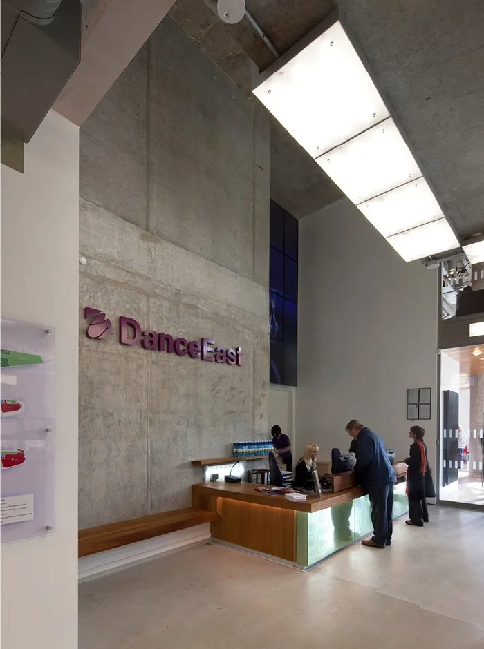 DanceEast reception area with tall concrete walls, a purple sign, and 2 people talking at the small reception counter.