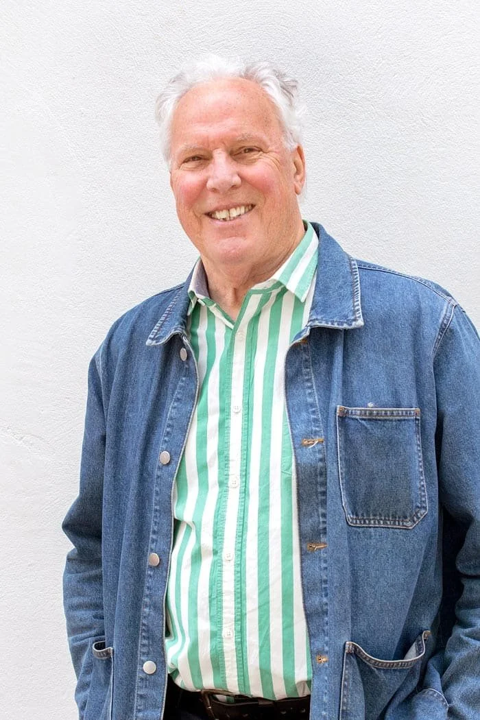 Smiling grey haired man wearing a denim jacket and a green and white striped shirt standing against a plain white wall.