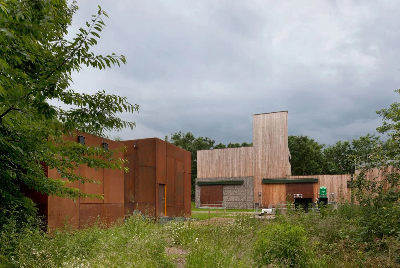 External view of Water Treatment Facility modern buildings with wooden and rust-coloured metal exteriors, surrounded by greenery under a cloudy sky with tree branches to give depth of field in the foreground.