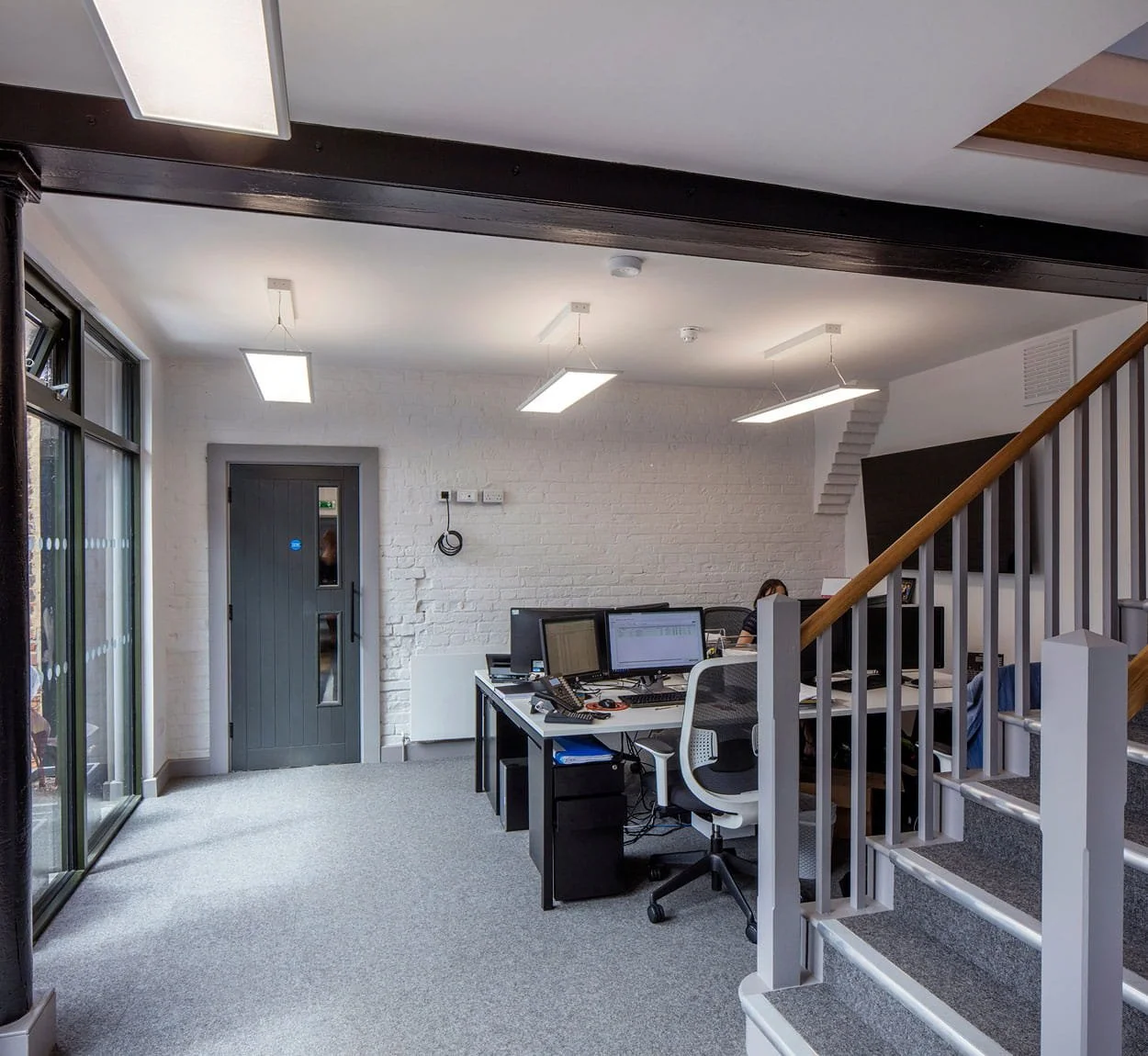 Office with multiple workstations, desktop computers, and office chairs, next to a staircase with a wooden handrail, white brick accent wall, and large windows letting in natural light.