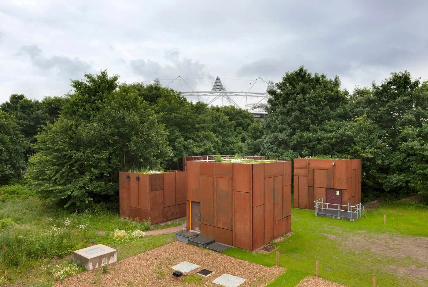 A raised view of three rust-coloured metal cube structures in a lush green park with trees and a cloudy sky in the background.