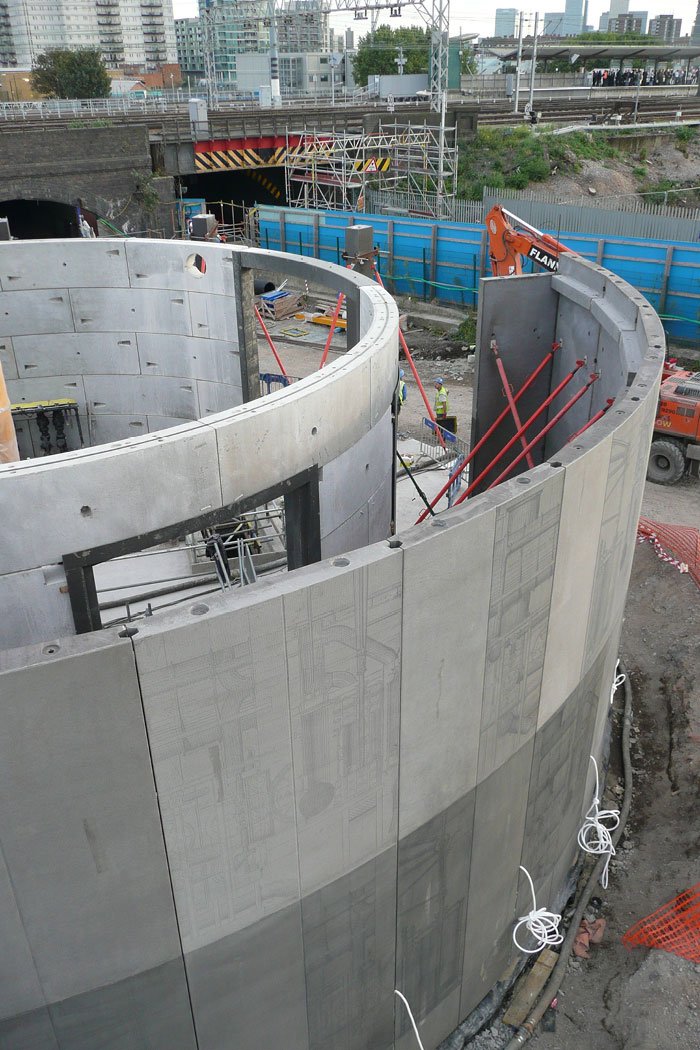 A close up of the construction site showing the curved-etched concrete wall panels being built – with scaffolding, construction workers, and heavy equipment on site with a railway bridge and city buildings in the background.
