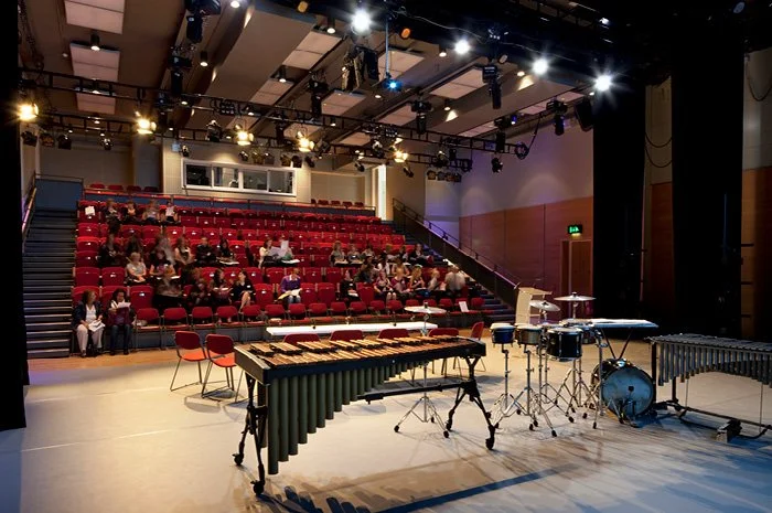 An indoor concert hall setting with the view from the back of an empty stage with musical instruments including a xylophone, drums, and cymbals, looking out to an audience seated in theatre seats.