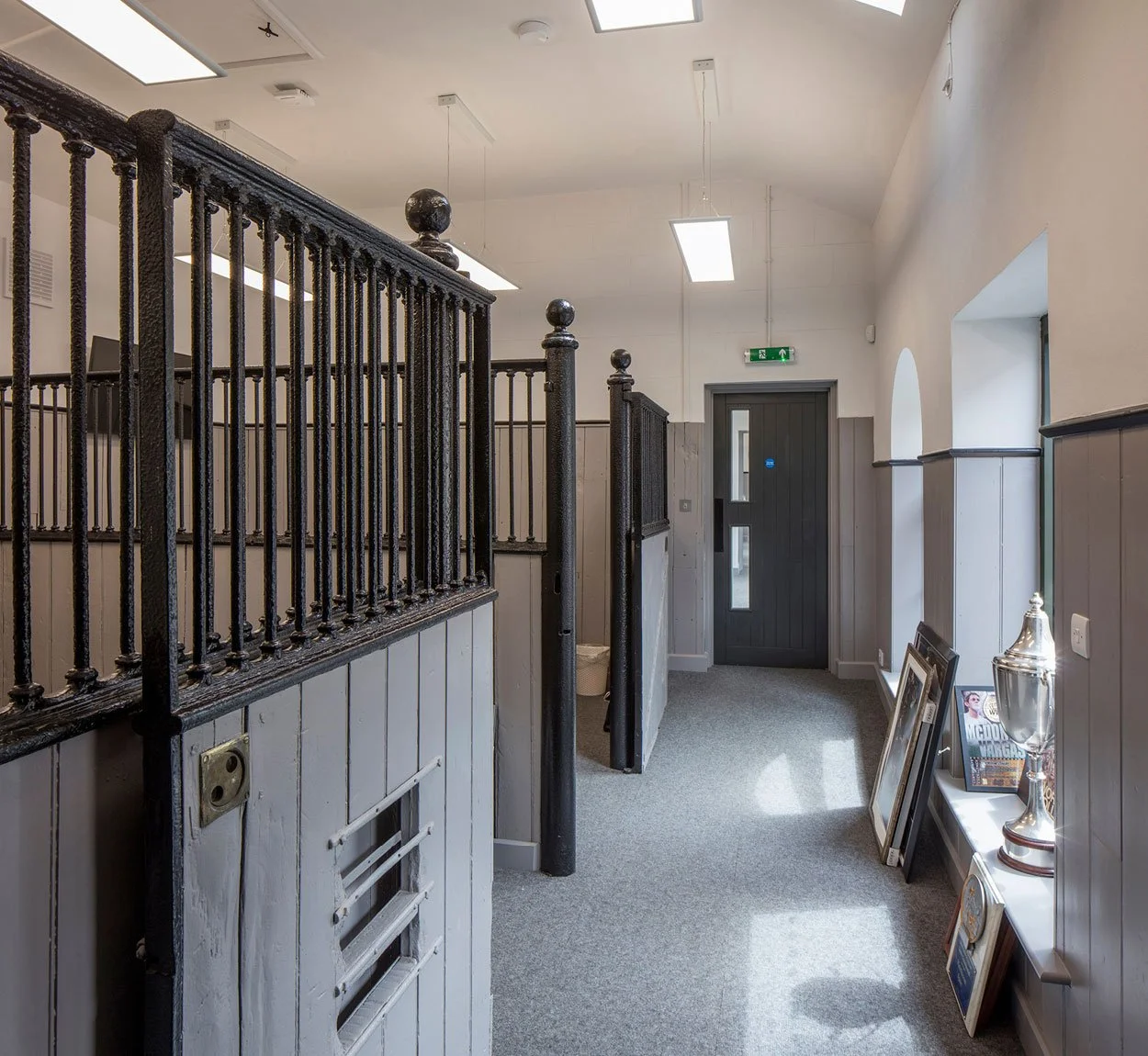 Interior hallway of a building with gray and white walls, black door, framed pictures, a decorative silver trophy, and a black metal railing.