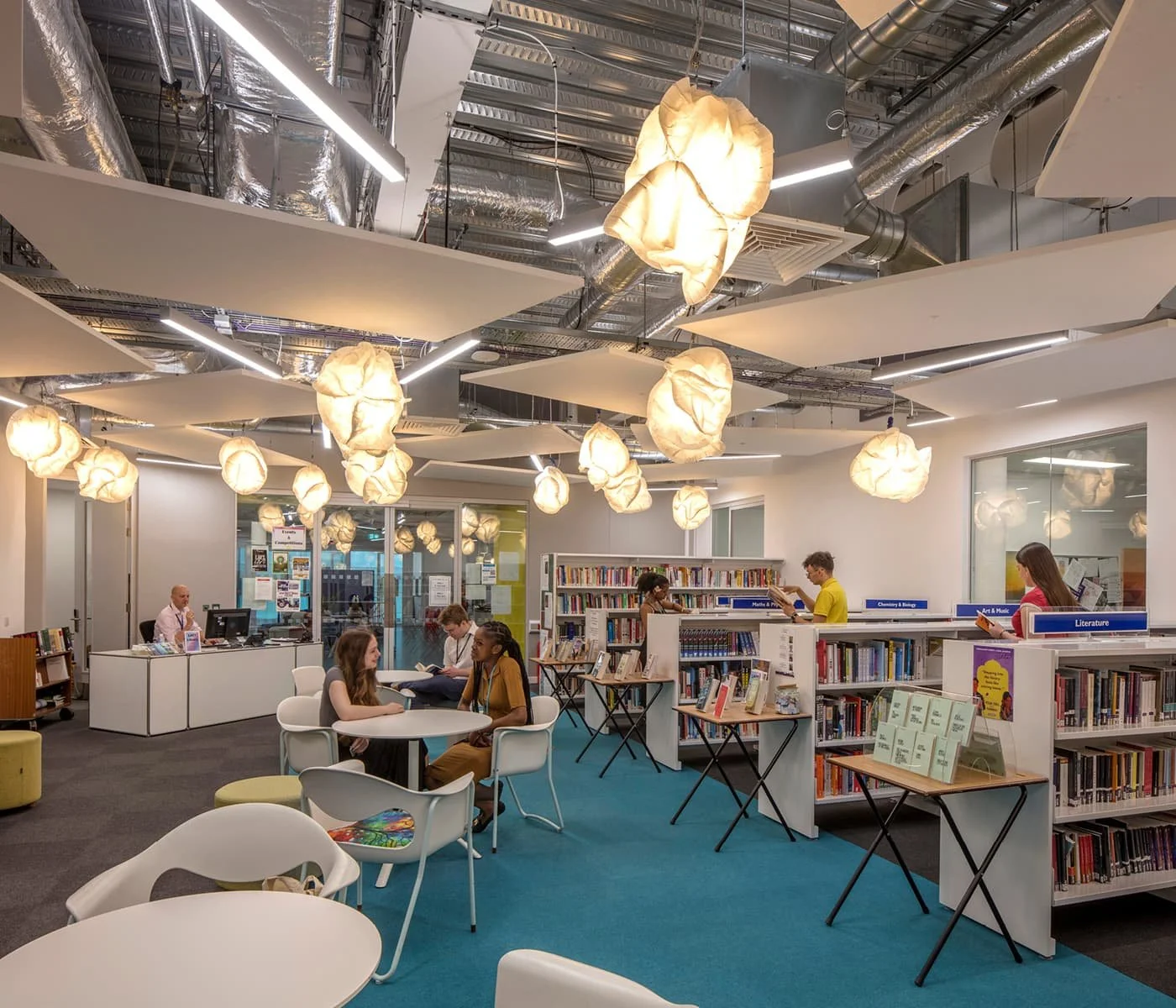 Interior view of a modern library with students studying and browsing books, ceiling with decorative lighting and exposed ductwork.