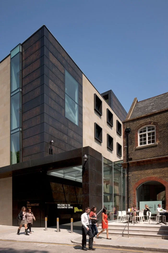 Modern building with glass and black panel facade, next to an older brick building, with people walking and sitting outside at a cafe.