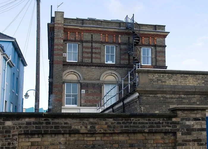 A distant rear view of the Tourist Information Centre's multi-story brick building with fire escape on the side, adjacent to a blue building and a similar historic-brick coloured wall in the foreground, taken from ground level.