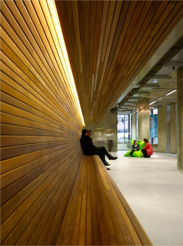 Interior space with a long, curved wooden bench against a wooden wall, with people sitting on the bench and colorful chairs nearby in a modern, industrial-style building.