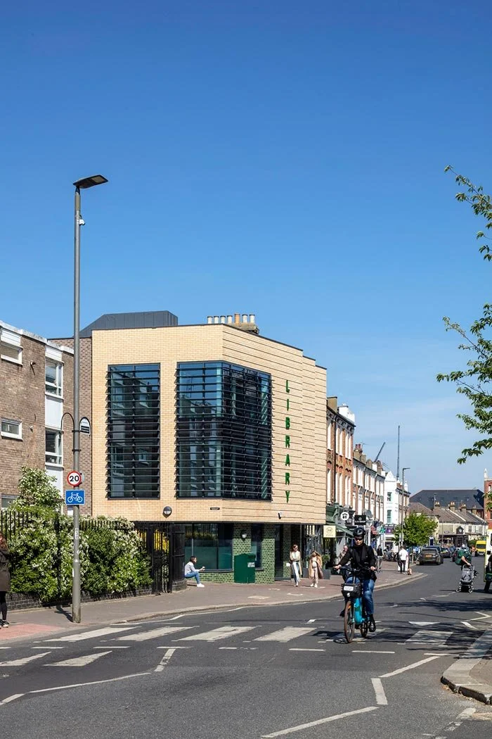 A street level view of the front entrance to Northcote Library building with a distinctive facade and the word 'LIBRARY' vertically on it, situated on a busy street with pedestrians and cyclists, under a clear blue sky.