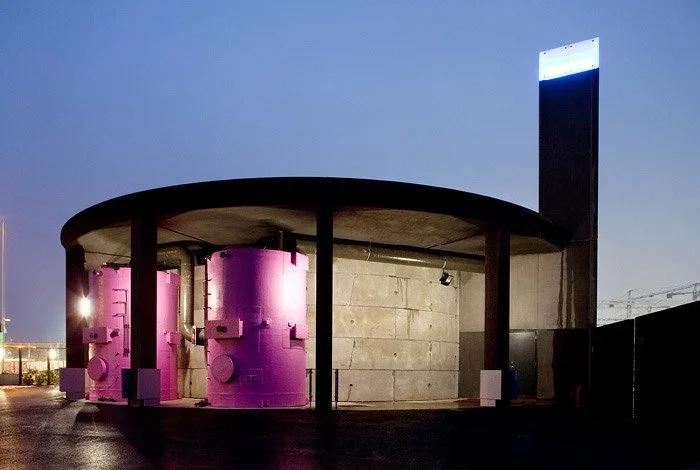 Modern building with cylindrical pink structures, a concrete wall, and a tall illuminated vertical sign against a clear evening sky.