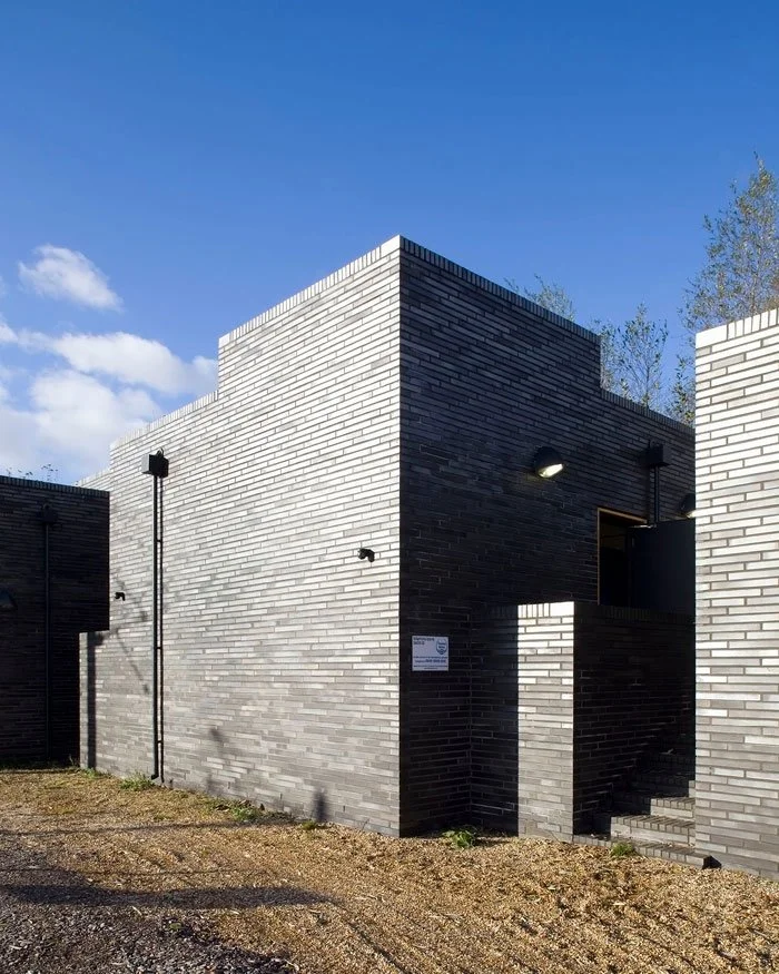 A close up of one of Stratford Box Pumping Station's modern building with dark grey brick walls, a small window, and outdoor lighting against a clear blue sky.