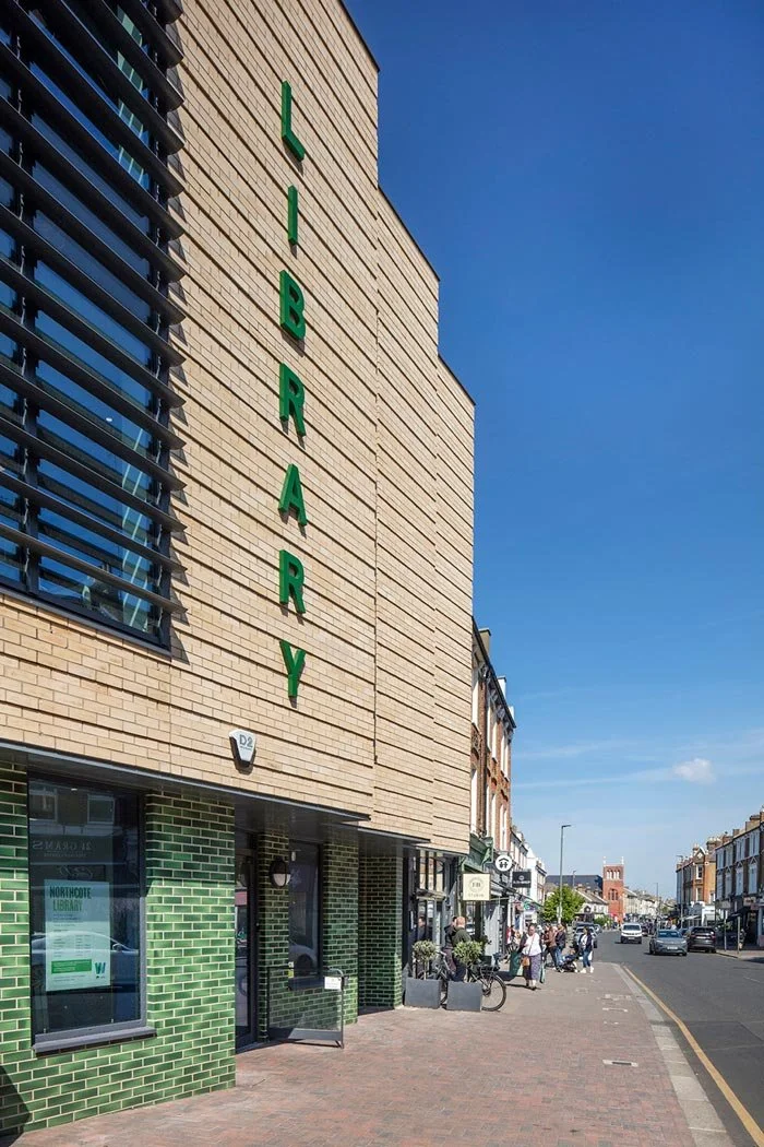 Closeup of the Northcote Library building with a large vertical green sign spelling 'LIBRARY' and a sidewalk with pedestrians, bicycles, and shops in the far distance.