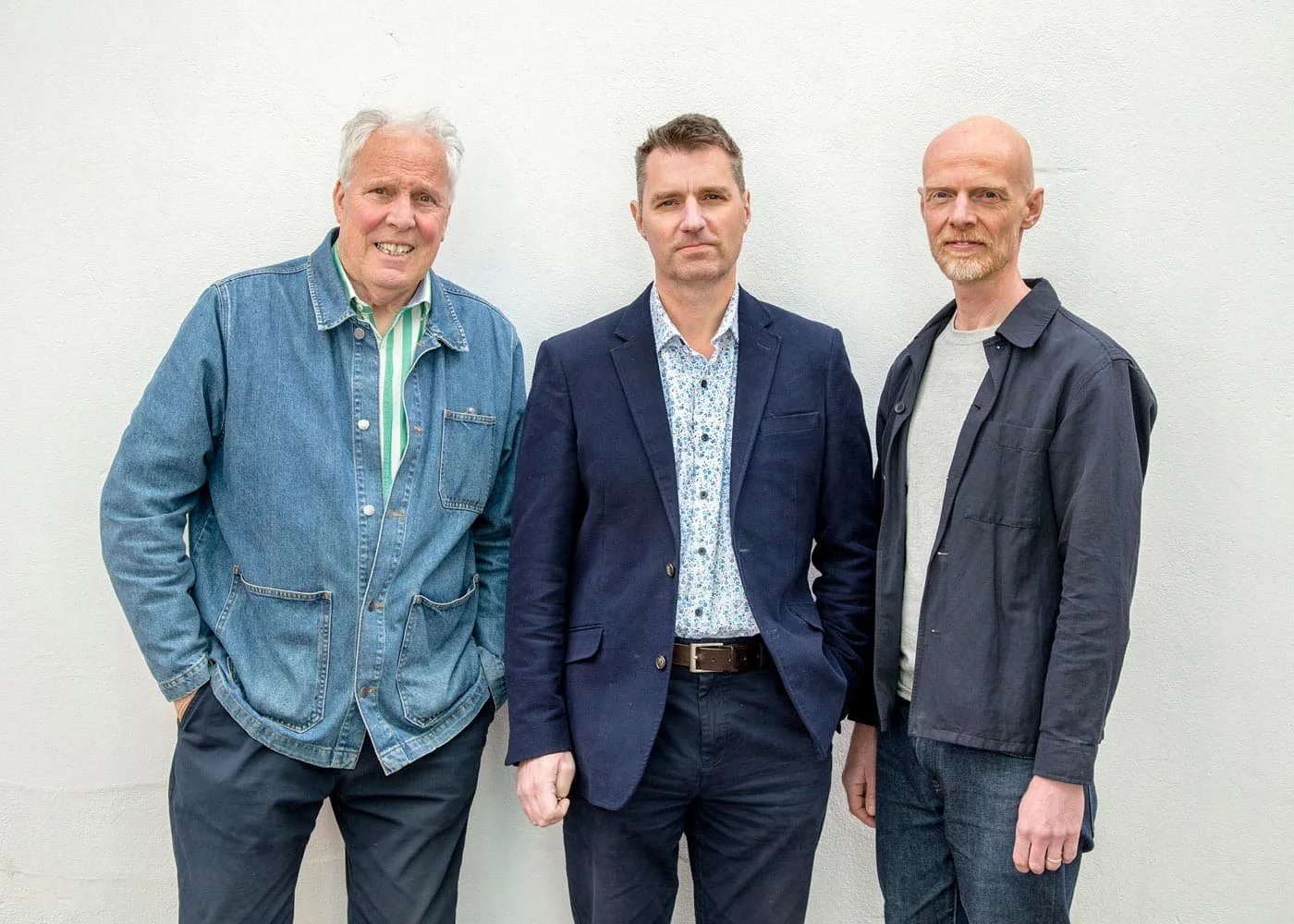 Three men standing side by side against a plain white wall, dressed in casual and semi-formal clothing.