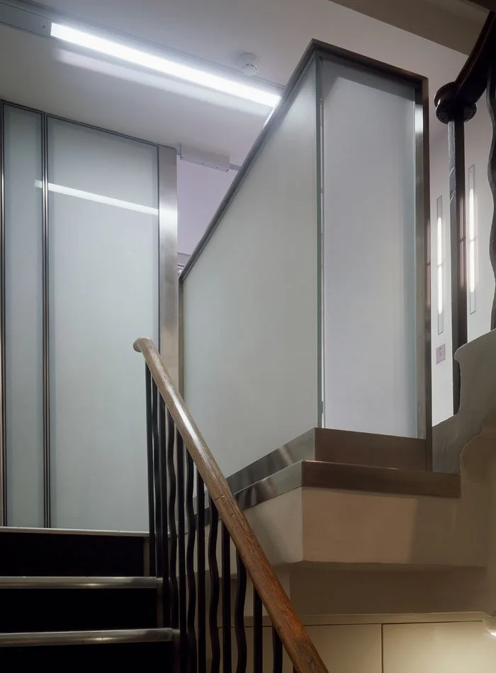 Interior view of Silver House staircase with black metal railings and wooden handrail, leading up to a frosted glass and metal partition wall in a building.