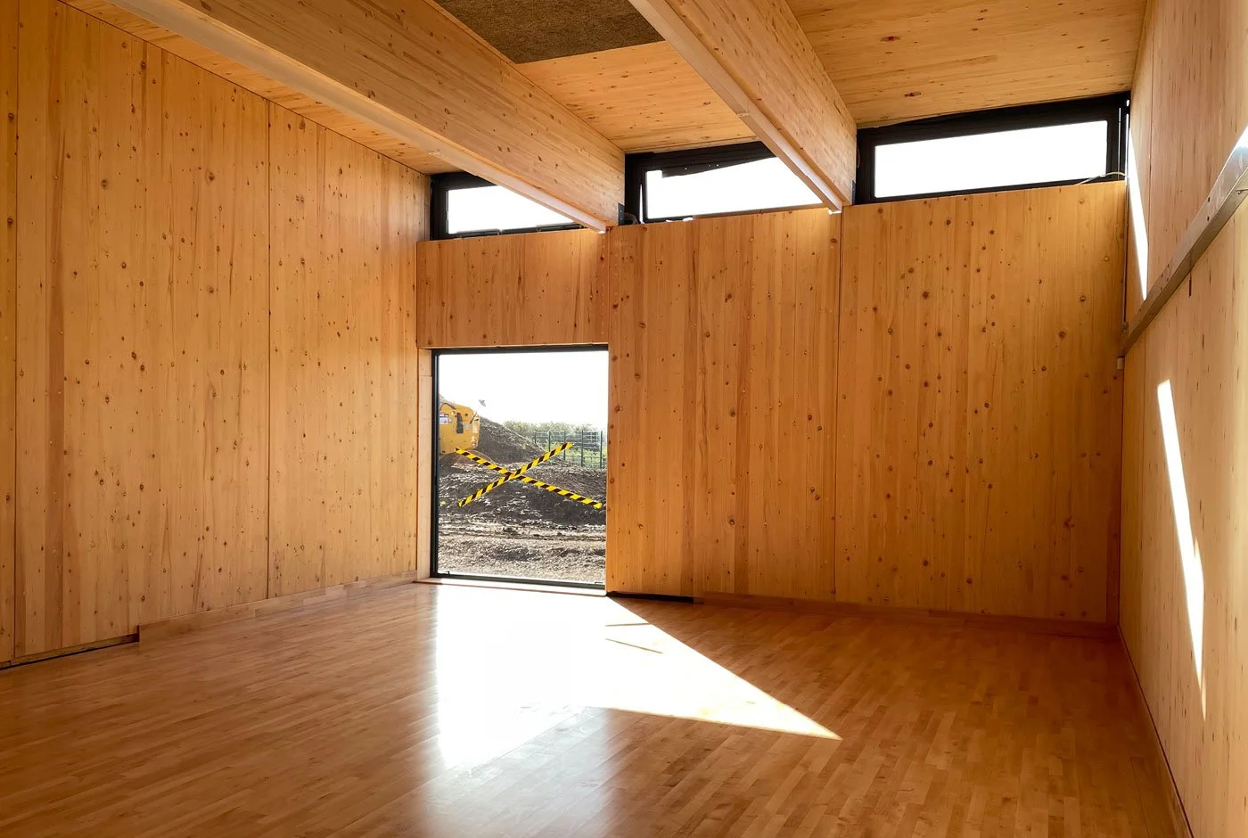Empty room with wooden walls, ceiling, and floor, illuminated by natural light from windows and door, with construction site outside.