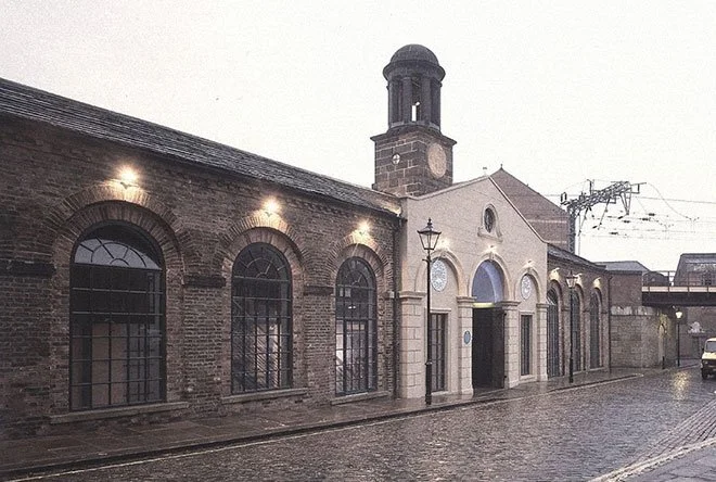 Historic brick building of White Cloth Hall in Leeds, with arched windows and a clock tower on a rainy, cobblestone street at dusk.