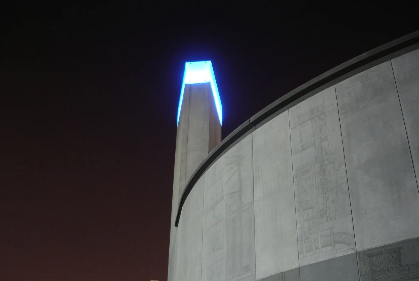 Night view The Pudding Mill Pumping Station with a closeup angle showing the tall tower illuminated with blue light in the centre of the image, with the curved exterior concrete panels filling the right foreground and the rest the night sky.