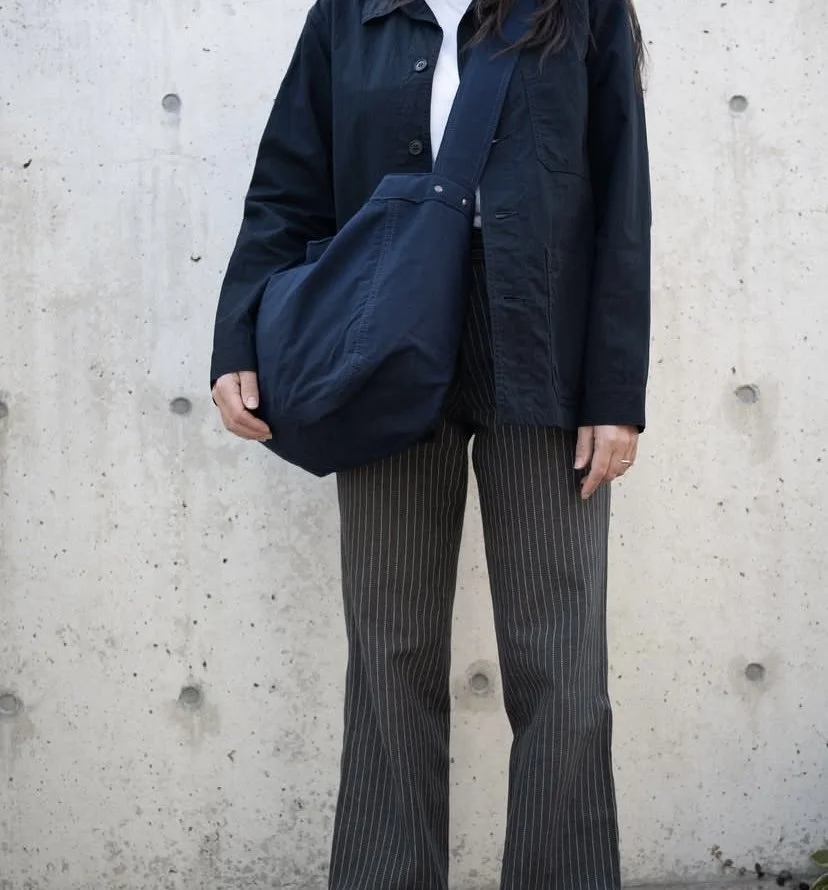 Person wearing a black jacket and pinstripe pants, carrying a large navy blue bag, standing against a concrete wall.
