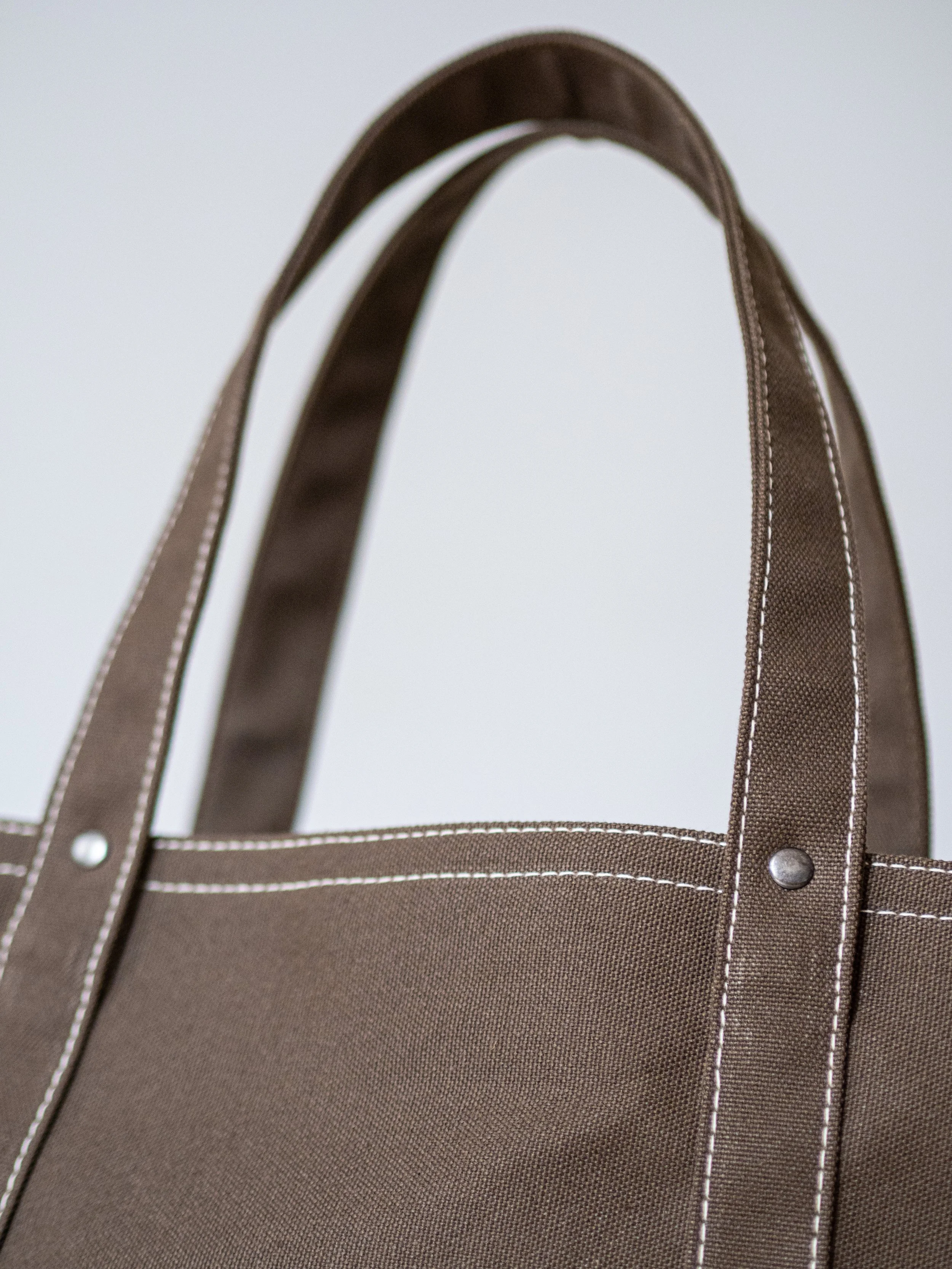 Close-up of a brown tote bag with white contrast stitching, showing handles and a metallic rivet.