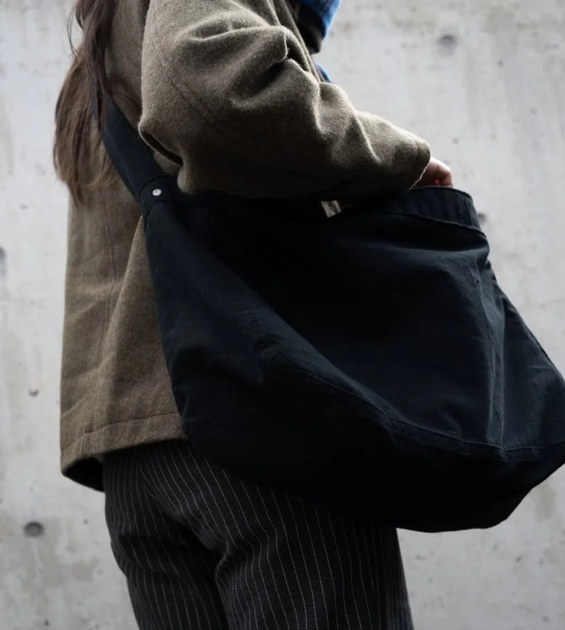 Person wearing a brown jacket and pinstriped pants holding a black vintage newspaper bag against a concrete wall.