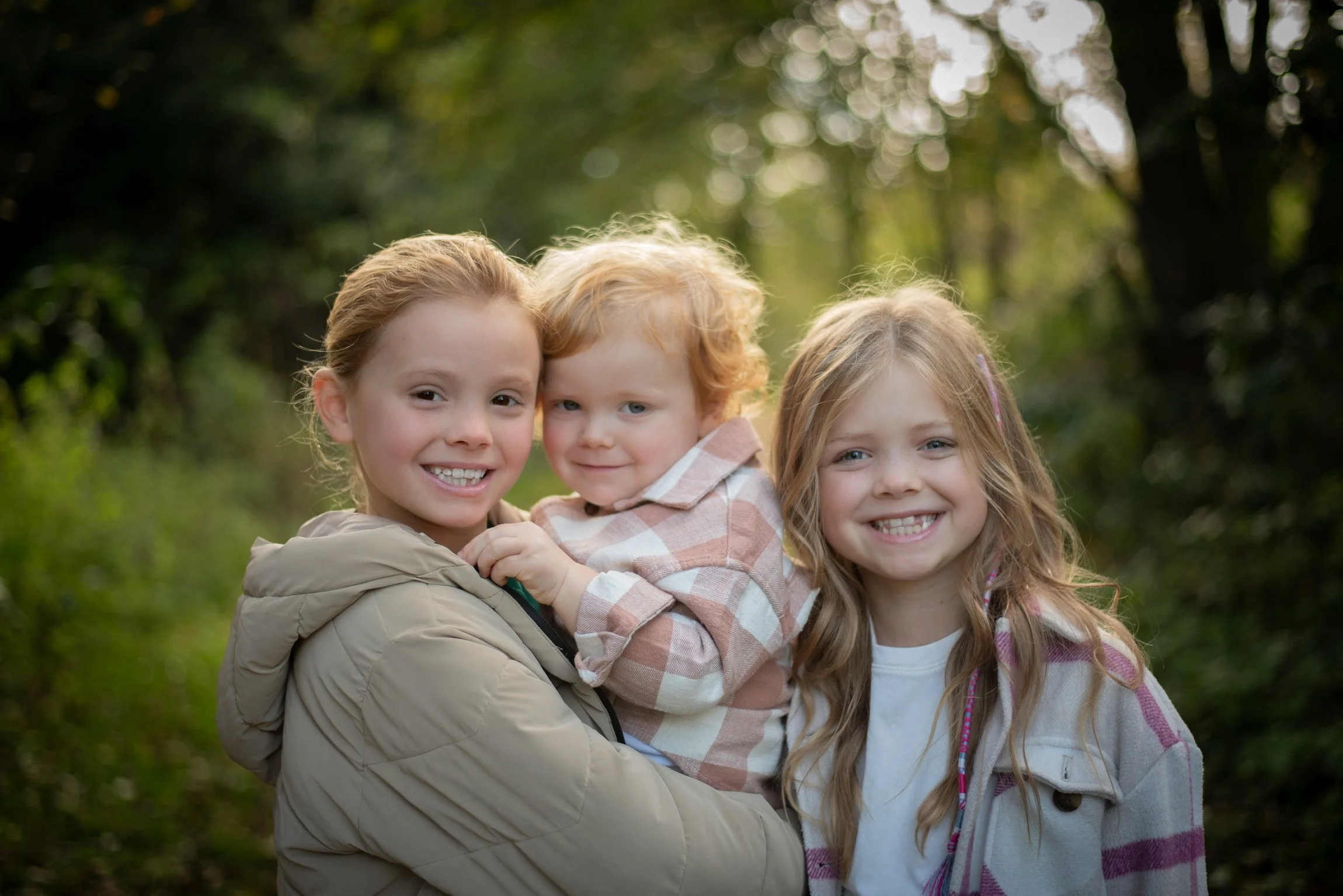 Three children smiling, outdoors in a wooded area, with sunlight filtering through trees.